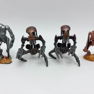 May include: A collection of four science fiction robot action figures. One silver humanoid droid, two brown multi-legged spider-like droids, and one slender reddish-brown bipedal droid, each standing on a textured base.