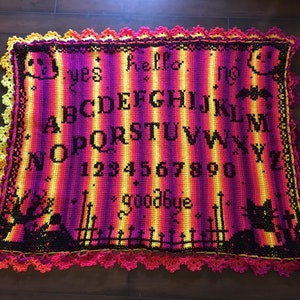 Könnte beinhalten: Eine gehäkelte Ouija-Brett-Decke in Lila-, Gelb- und Orangetönen. Die Decke zeigt das Alphabet, Zahlen und die Wörter "Ja", "Nein", "Hallo" und "Auf Wiedersehen". Die Ränder sind mit einer dekorativen Häkelborte versehen.
