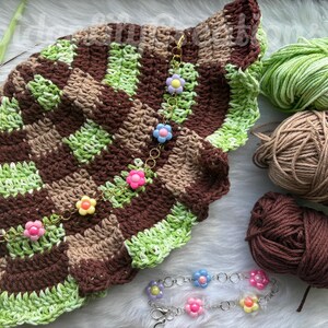 Peut inclure: Un chapeau crocheté à carreaux marron et vert, orné d'une chaîne de breloques fleuries. Plusieurs pelotes de laine vertes, marron et beige sont à côté du chapeau. Crochet et bracelet de fleurs.
