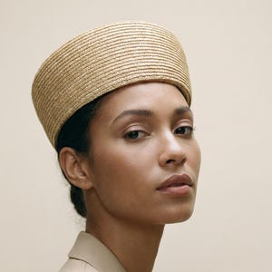 May include: A woman wearing a tan blazer and a straw boater hat. The hat is a light beige colour and has a flat top and a rounded brim. The blazer is a neutral tan colour and has a classic tailored fit. The background is a solid, light beige colour.