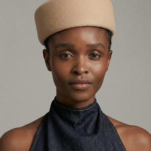 May include: A tan-colored, pillbox-style hat. The hat has a flat top and a rounded, cylindrical shape. The hat is made of a soft, felt-like material. The model is wearing a dark blue denim halter top.