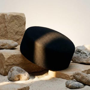 May include: A black, rounded hat with a flat top, placed on a collection of tan and beige rocks and sand. The hat is the central object, with a shadow across its surface. The scene is illuminated by natural light.