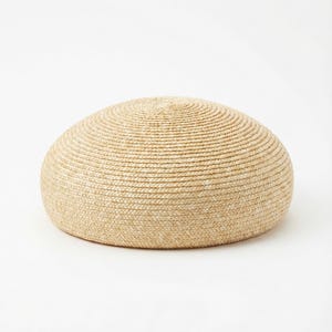 Könnte beinhalten: Ein runder, gewebter Hocker in Naturbeige. Der Hocker hat eine gewölbte Form und besteht aus dicht gewebtem, strukturiertem Material. Das Design weist konzentrische Kreise auf, die ein optisch ansprechendes Muster erzeugen. Die neutrale Farbe macht ihn für verschiedene Einrichtungsstile geeignet.