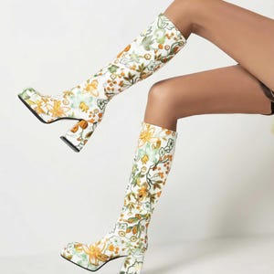 May include: White knee-high boots with a floral pattern in shades of yellow, orange, and green. The boots have a chunky heel and platform sole. The floral design covers the entire boot, creating a bold and stylish look.