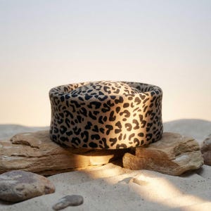 May include: A round, leopard print hat with a wide brim. The hat is made of a soft, textured fabric and features a repeating pattern of black spots on a tan background. The hat is displayed on a rock.