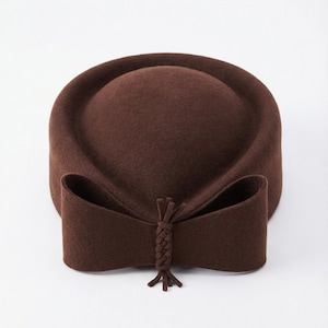May include: A brown felt hat with a rounded crown and a wide, decorative bow at the front. The bow has a braided detail in the centre and a fringe. The hat is a solid, warm brown colour.