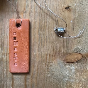 Op de afbeelding: Een terracotta-label met het woord "Namaste" erop. Het label is bevestigd aan een touwtje.