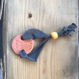 Könnte beinhalten: Ein braunes Ton-Etikett mit dem Text "CAT MOM" und einem Pfotenabdruck. Das Etikett ist an einem grauen Stoffband mit einer gelben Holzperle befestigt.