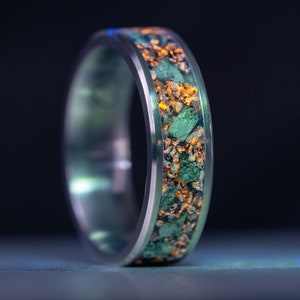 Puede incluir: Un anillo plateado con una banda llena de piedras verdes y anaranjadas. El anillo está sobre un fondo oscuro, con una luz azul que ilumina la parte inferior. El diseño del anillo es moderno y llamativo.