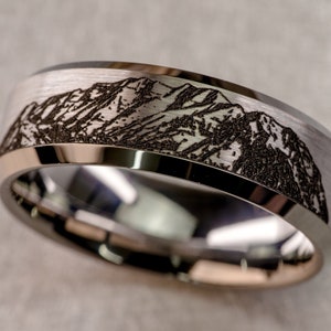Op de afbeelding: Een zilveren trouwring met een geborstelde afwerking en een gegraveerd berglandschap.