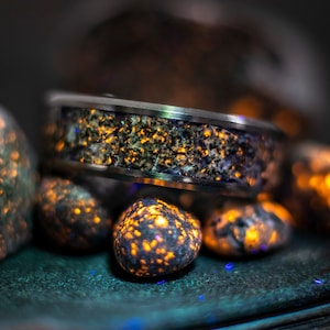Può includere: Un anello d'argento con un intarsio scuro e macchiettato, appoggiato tra diverse piccole pietre luminose. Le pietre e l'intarsio dell'anello presentano riflessi arancioni e blu, creando un contrasto sorprendente sullo sfondo scuro. L'anello è il punto focale.