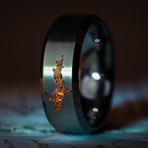 Puede incluir: Un anillo de carburo de tungsteno plateado con un diseño recortado de un lago en el centro. El lago está lleno de un material naranja brillante.