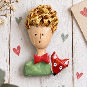 May include: A sculpted brooch featuring a figure with textured golden-brown hair, a tan face, a bright red bow tie, and a green shirt. A small red fox with white eyes and a black nose is positioned on the right shoulder of the figure.
