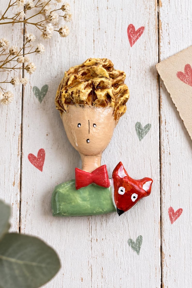May include: A sculpted brooch featuring a figure with textured golden-brown hair, a tan face, a bright red bow tie, and a green shirt. A small red fox with white eyes and a black nose is positioned on the right shoulder of the figure.