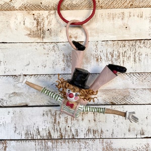 Puede incluir: Un muñeco de payaso de papel maché hecho a mano con pelo castaño rizado, nariz roja y camisa a rayas verde y blanca. El payaso está colgado de dos aros rojos y rosas, con los brazos extendidos.