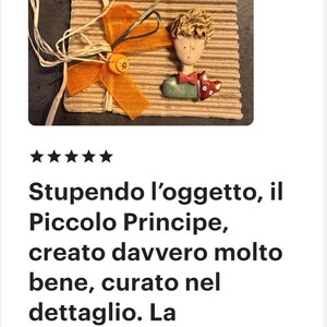 May include: A decorative wall hanging featuring a sculpted figure of a boy with blonde hair and a red bowtie, accompanied by a small fox figurine. The piece is mounted on a rectangular piece of corrugated cardboard with an orange ribbon and button.