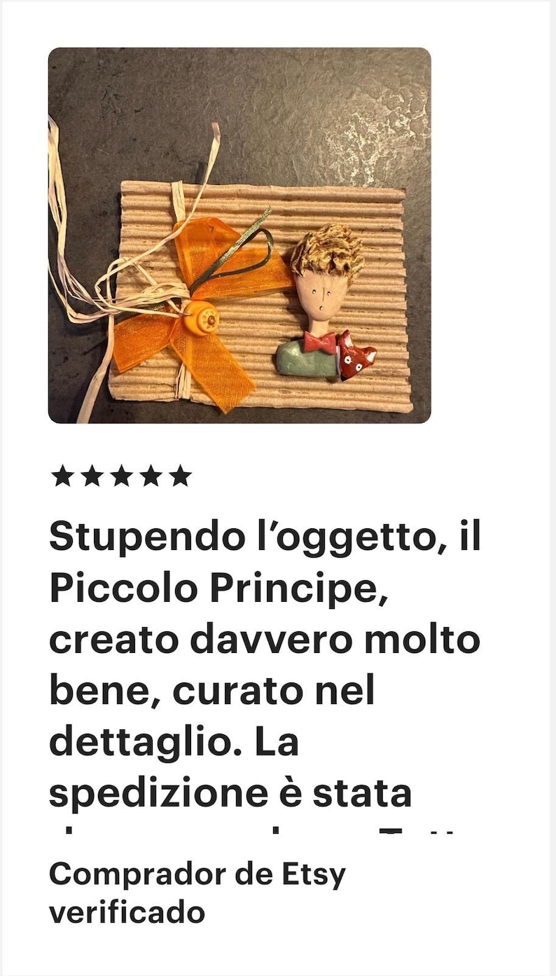 May include: A decorative wall hanging featuring a sculpted figure of a boy with blonde hair and a red bowtie, accompanied by a small fox figurine. The piece is mounted on a rectangular piece of corrugated cardboard with an orange ribbon and button.
