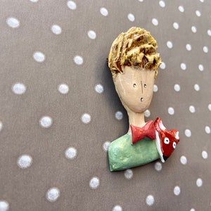 May include: A sculpted brooch featuring a figure with textured blonde hair, a pale face, a green shirt, and a red bow tie. A small red and white fox is positioned on the shoulder of the figure. The piece has a smooth, glossy finish throughout.