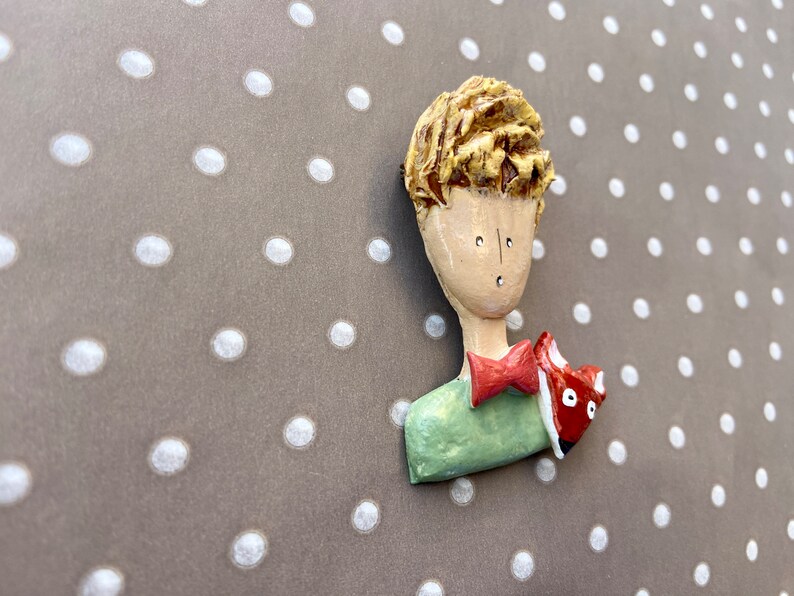 May include: A sculpted brooch featuring a figure with textured blonde hair, a pale face, a green shirt, and a red bow tie. A small red and white fox is positioned on the shoulder of the figure. The piece has a smooth, glossy finish throughout.