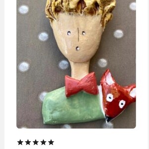 May include: A sculpted brooch featuring a figure with textured yellow hair, a pale face, and a small mouth. The figure wears a green shirt with a red bow tie and is accompanied by a small red fox with white eyes positioned on the right shoulder.