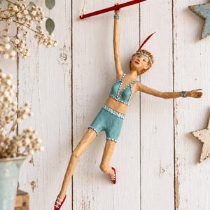 Puede incluir: Una figura decorativa de un acróbata colgado de una barra de trapecio roja. El personaje viste un conjunto de dos piezas azul claro con adornos de lentejuelas plateadas, una diadema a juego y zapatos rojos, con un accesorio de pluma roja.