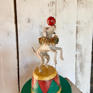 May include: A white paper mache dog balancing a red ball on its nose. The dog is wearing a green and gold bow and is standing on a yellow, red, and green paper mache circus platform.
