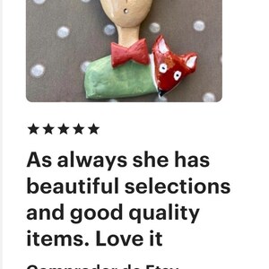 May include: A handcrafted brooch featuring a stylized figure with textured golden-brown hair, a pale face, a red bow tie, and a light green shirt. A small red fox with white accents is positioned next to the figure, resting against the green shirt.
