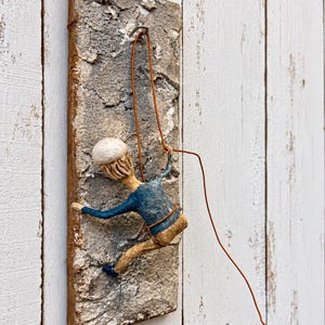 Puede incluir: Una escultura de pared que muestra una figura escalando un panel rectangular gris texturizado. La figura viste una camiseta azul y pantalones color canela, con una gorra blanca. Un alambre fino y flexible de color cobre se extiende.