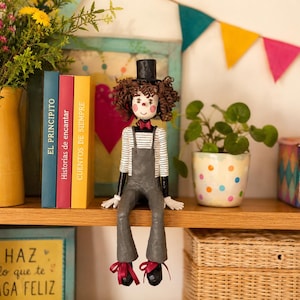 Puede incluir: Una muñeca artesanal con cabello castaño rizado, un sombrero de copa negro y una pajarita roja. La muñeca viste una camisa blanca de manga larga con finas rayas horizontales negras bajo un peto gris, guantes blancos y zapatos negros.