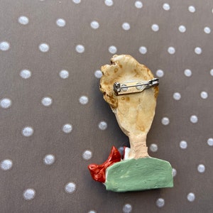May include: A brooch featuring a sculpted figure with light brown hair, a green shirt, and a red scarf. The back of the piece is a light tan color and includes a silver-toned metal pin mechanism with a rotating clasp for secure attachment.