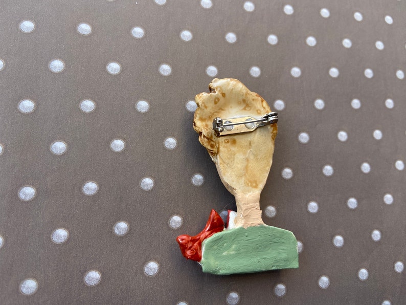 May include: A brooch featuring a sculpted figure with light brown hair, a green shirt, and a red scarf. The back of the piece is a light tan color and includes a silver-toned metal pin mechanism with a rotating clasp for secure attachment.
