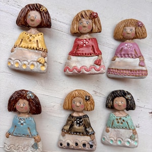 Puede incluir: Seis broches de cerámica con forma de muñeca y detalles pintados. Cada figura presenta un color de pelo único, una parte superior de color con detalles florales y una falda blanca con dobladillo festoneado. Las muñecas tienen mejillas rosadas.