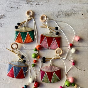 May include: A selection of colourful, handcrafted drum ornaments. Each ornament features a painted drum design with a wooden ring, drumsticks, and a string of beads and bells. The drums are painted in various colours, including red, green, and blue.