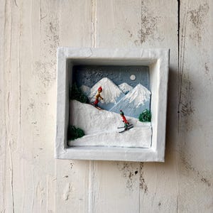 Peut inclure: Diorama d'hiver encadré représentant une scène de montagne enneigée avec deux skieurs. Les figurines miniatures portent des tenues rouges et jaunes, skiant sur une pente enneigée. L'arrière-plan comprend des montagnes enneigées, une pleine lune et des arbres.
