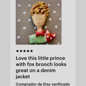 May include: A handcrafted brooch featuring a figure with textured golden-brown hair, a pale face, a red bow tie, and a green shirt. A small red and white fox is positioned next to the figure. The brooch has a glossy finish and detailed features.