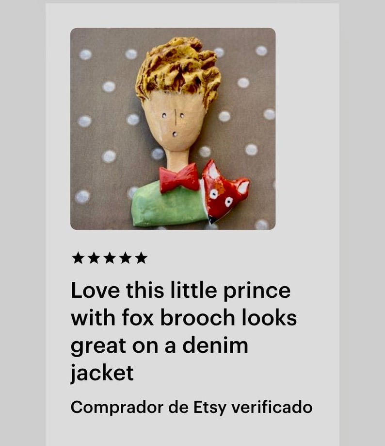 May include: A handcrafted brooch featuring a figure with textured golden-brown hair, a pale face, a red bow tie, and a green shirt. A small red and white fox is positioned next to the figure. The brooch has a glossy finish and detailed features.