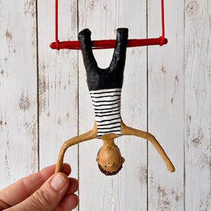 Puede incluir: Una figura de una persona colgada boca abajo de una barra de trapecio roja. La figura tiene cabello castaño, piel bronceada y viste una camiseta de rayas horizontales blancas y negras con pantalones negros. El trapecio cuelga de dos cuerdas rojas.