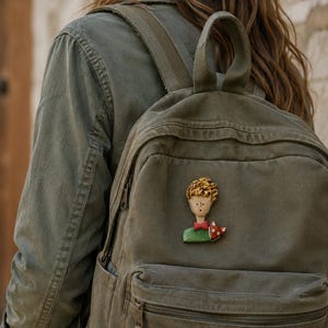May include: A small, hand-painted brooch featuring a stylized character with blonde hair, a red bowtie, and a green shirt, accompanied by a small orange fox. The brooch is attached to the front pocket of an olive green canvas backpack.