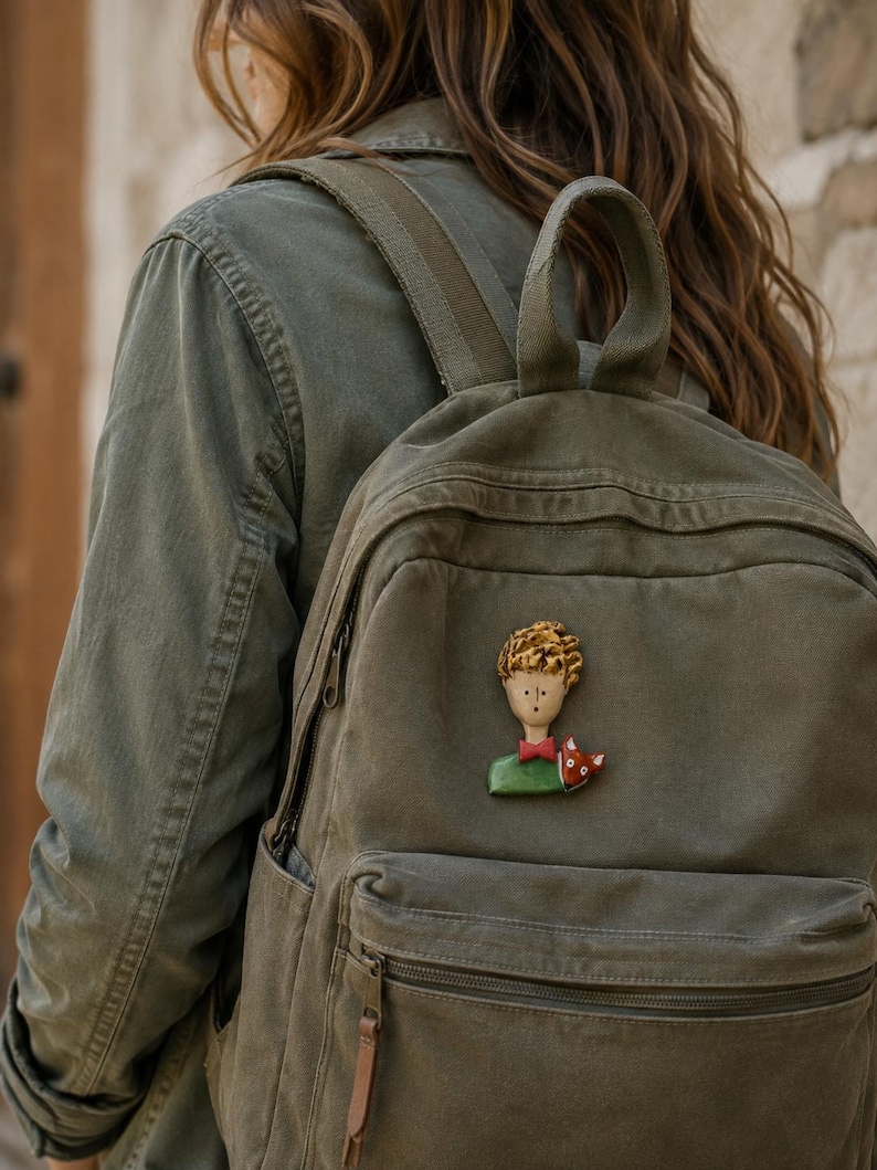 May include: A small, hand-painted brooch featuring a stylized character with blonde hair, a red bowtie, and a green shirt, accompanied by a small orange fox. The brooch is attached to the front pocket of an olive green canvas backpack.