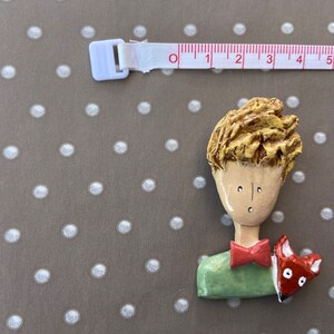 May include: A hand-painted brooch depicting a character with textured golden hair, a light tan face, a green shirt, and a red bowtie, accompanied by a small orange fox. A white measuring tape shows the figure is approximately 2.5 inches wide.