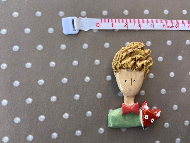 May include: A hand-painted brooch depicting a character with textured golden hair, a light tan face, a green shirt, and a red bowtie, accompanied by a small orange fox. A white measuring tape shows the figure is approximately 2.5 inches wide.