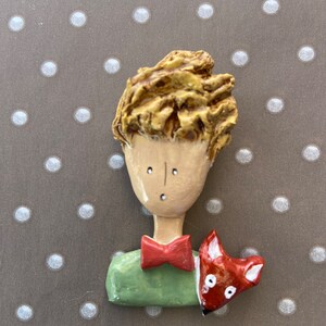 May include: A sculpted brooch featuring a figure with textured golden-brown hair, a tan face, a red bow tie, and a green shirt. A small red and white fox is positioned on the figure's shoulder. The piece has a smooth, glossy finish throughout.