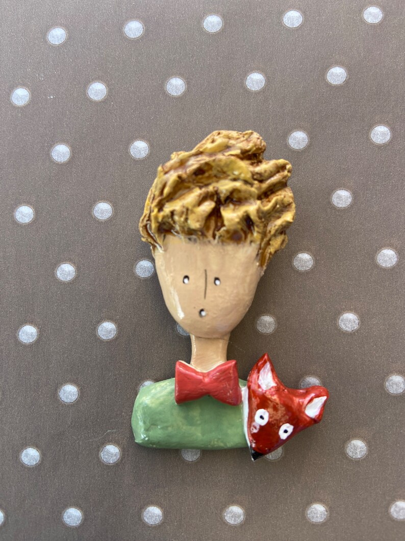 May include: A sculpted brooch featuring a figure with textured golden-brown hair, a tan face, a red bow tie, and a green shirt. A small red and white fox is positioned on the figure's shoulder. The piece has a smooth, glossy finish throughout.