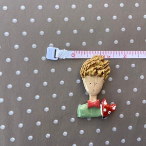 May include: A sculpted brooch depicting a person with textured blonde hair, a red bowtie, and a green shirt, accompanied by a small red fox. A white measuring tape is positioned above the brooch, showing a width of approximately 2.5 inches.