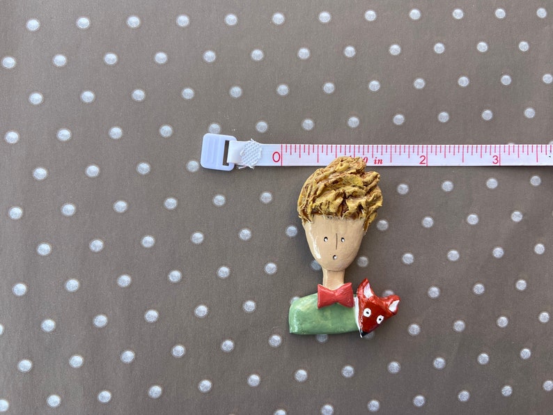 May include: A sculpted brooch depicting a person with textured blonde hair, a red bowtie, and a green shirt, accompanied by a small red fox. A white measuring tape is positioned above the brooch, showing a width of approximately 2.5 inches.
