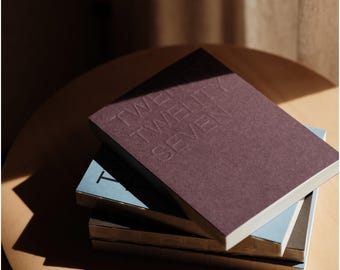May include: A stack of four rectangular notebooks with exposed stitched spines. The top notebook is dark purple with the embossed text TWENTY TWENTY SEVEN. The notebooks below are light blue, dark gray, and white, featuring visible binding.