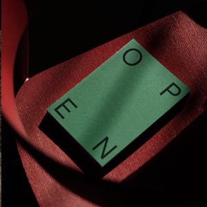 May include: A rectangular green card with the word OPEN printed in black capital letters, positioned diagonally across a textured dark red surface. The card features a matte finish and clean, sharp edges, contrasting with the deep red background.