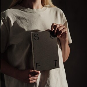 May include: A rectangular notebook with a dark gray cover featuring the letters S, O, F, and T printed in black. The notebook has a visible stitched spine binding and is held against a plain, light-colored short-sleeved t-shirt.