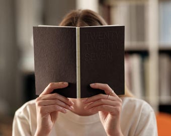 May include: A dark brown notebook with a visible white spine binding. The front cover features the embossed text TWENTY TWENTY SEVEN. The notebook is held open, displaying a textured paper surface and a clean, minimalist design aesthetic.