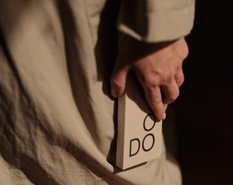 May include: A rectangular beige box with the black text DO printed on the front surface. The box is held against a beige fabric garment. The lighting creates shadows across the surface of the box and the fabric, highlighting the texture.